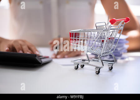 Close-up Of Person Calculating Bill With Calculator Near Shopping Cart On Desk Stock Photo