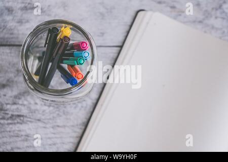 Overhead shot of colorful markers in a jar next to an opened textbook on a wooden surface Stock Photo