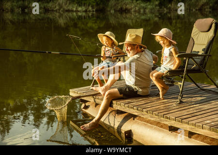 Cute little girls and their granddad are on fishing at the lake or river. Resting on pier near by water and forest in sunset time of summer day. Concept of family, recreation, childhood, nature. Stock Photo