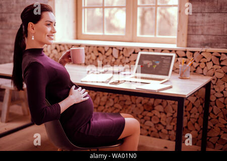 Healthy pregnancy. Kind woman keeping smile on her face while sitting in semi position Stock Photo