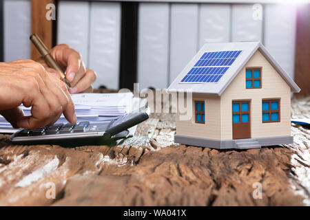 Businessman Using Calculator By Model Home With Solar Panel On Wooden Table Stock Photo
