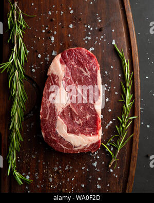 Raw ribeye steak with fresh rosemary on wooden cutting board, top down view. Vertical orientation Stock Photo