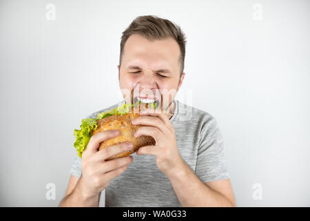 young handsome hungry man with closed eyes biting fresh sandwich with salad leaf on isolated white background. Stock Photo