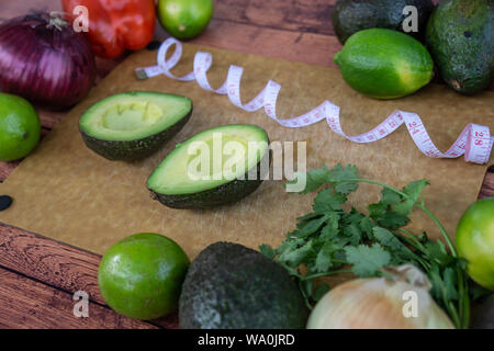 Halved avocado guacamole ingredients on wooden cutting board with measuring tape. Fresh cilantro, onion, limes and red bell pepper. Healthy raw diet f Stock Photo