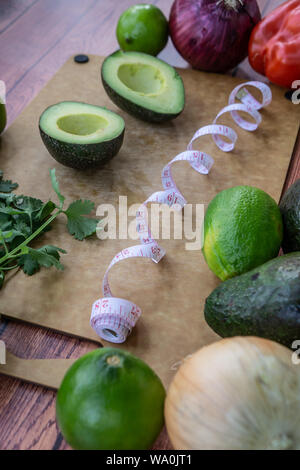 Halved avocado guacamole ingredients on wooden cutting board with measuring tape. Fresh cilantro, onion, limes and red bell pepper. Healthy raw diet f Stock Photo