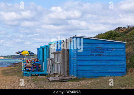 Mature couple sitting under parasol reading outside beach huts at Barton-on-Sea on a warm sunny day at Barton on Sea, Hampshire, UK in August Stock Photo