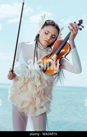 tender woman in white swan costume standing on blue sky background with violin Stock Photo