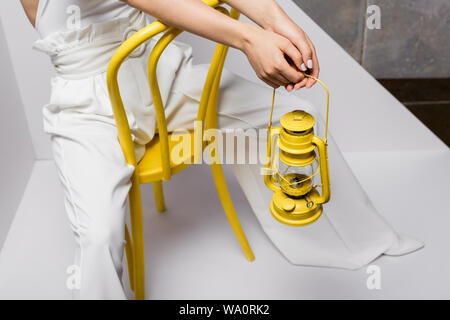 cropped view of young woman sitting on yellow chair and holding retro lamp on white and grey Stock Photo