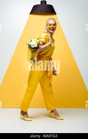 cheerful blonde woman holding string bag with flowers while posing on white and yellow Stock Photo
