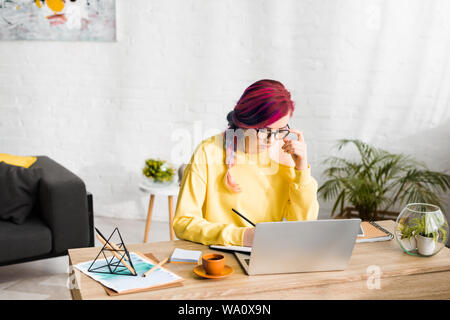 hipster girl pensively making notes while sitting behind table in living room Stock Photo