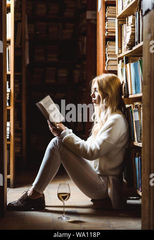 side view of blonde woman reading book and sitting on floor in library Stock Photo