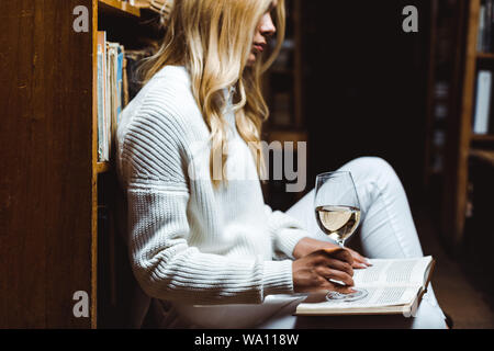 side view of blonde woman reading book and holding glass wine in library Stock Photo