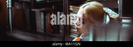 panoramic shot of blonde and attractive woman in glasses in library Stock Photo