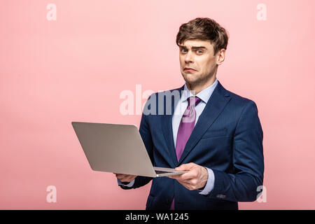 disappointed businessman looking at camera while holding laptop on pink background Stock Photo