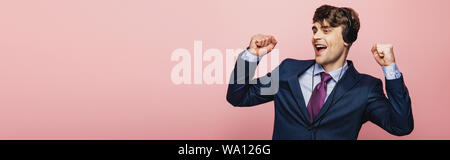 panoramic shot of excited businessman dancing while listening music in earphones on pink background Stock Photo