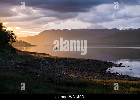 Sun rising over distant village of Lochcarron, with single boat silhouetted on a calm sea loch. Stock Photo