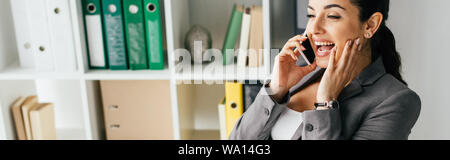 panoramic shot of pregnant woman sitting behind table in office and talking on smartphone Stock Photo