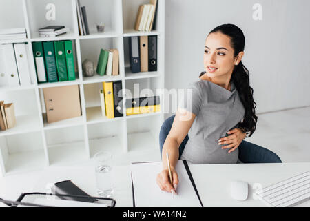 pregnant woman taking notes while sitting behind table in office and looking away Stock Photo