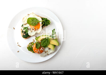 Fried eggs on a toasts bread with avocado, spinach and seeds on a white plate on the white background Stock Photo