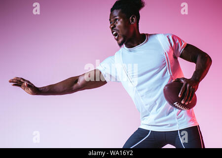 excited african american sportsman playing american football on purple background with gradient Stock Photo