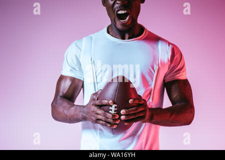 cropped view of african american sportsman yelling while holding rugby ball on purple background with gradient Stock Photo