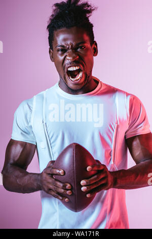 excited african american sportsman screaming at camera while holding rugby ball on purple background with gradient Stock Photo