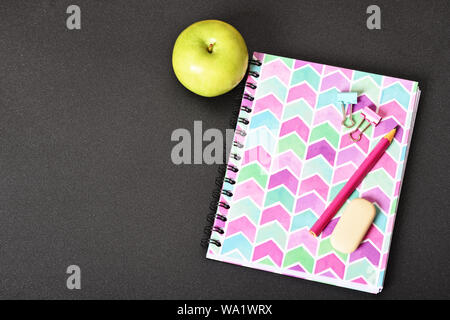 Top view of girly pink notebook with pencil, eraser, binder clips and apple on black background. Stock Photo