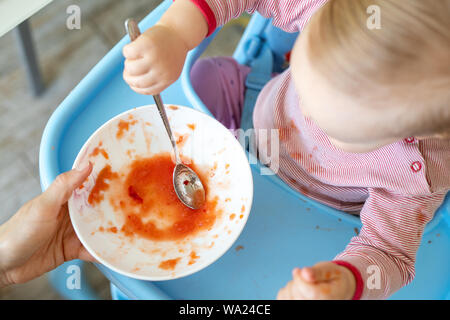 Adorable cute caucasian blond toddler boy eating tasty vegetable soup in chair at kithcen indoor. Mother feeding baby healthy food with spoon. Natural Stock Photo