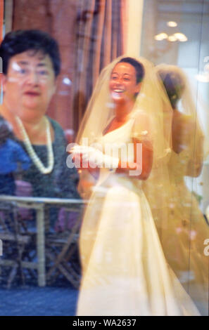 In a bridal shop, a young bride to-to-be smiles as she is being fitted into her wedding gown, as her mother looks on. Stock Photo