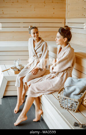 Two young beautiful girlfriends in bathrobes sitting together while relaxing in the luxury sauna Stock Photo