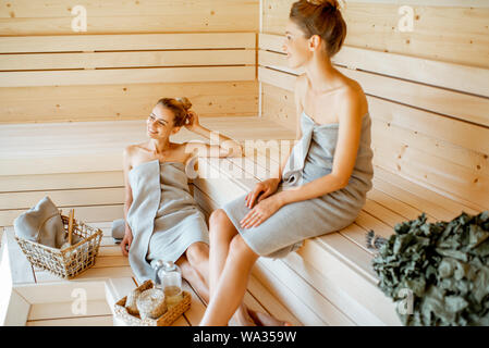Two young girlfriends wrapped in sheets relaxing in the sauna. Concept of female friendship and spa treatment Stock Photo