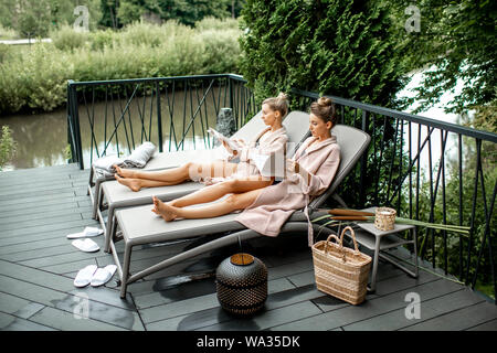 Two young women in bathrobes lying on the sunbeds, relaxing and spending time at the luxury SPA outdoors on the terrace