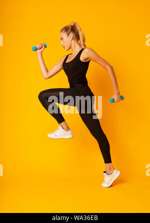 Young Woman Exercising Jumping With Dumbbells On Yellow Studio Background Stock Photo