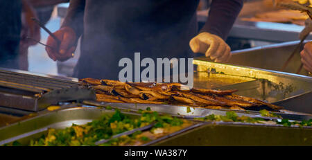 Chef with a pair of pliers and rubber gloves prepares fresh mackerel fillets for street sale on the grill at the sales stand Stock Photo
