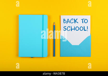 Back to school handwritten sign on white paper in blue envelope next to leather cover notebook over yellow paper background with copy space Stock Photo