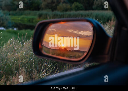 A car´s outside rear view mirror showing a golden sunset over green fields behind in a lush meadow Stock Photo