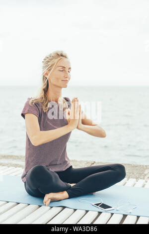 A Caucasian blonde woman in lotus position greets the namaste before starting yoga practice. Stock Photo