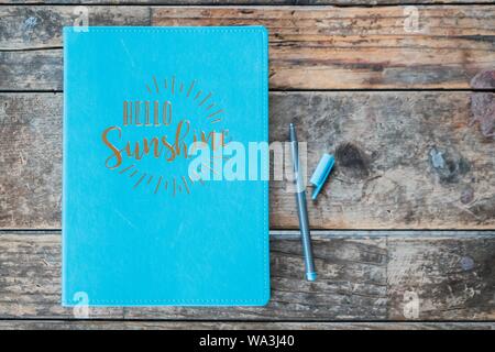 Overhead shot of a "hello sunshine" blue textbook with a ball-point pen next to it on wooden surface Stock Photo
