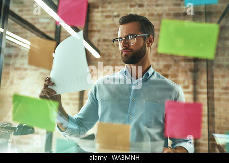 Creativity. Young and handsome bearded man in eyeglasses planning working process and using colorful stickers while standing in front of glass wall. Project management Stock Photo