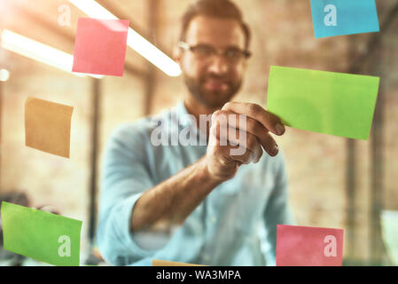 Kanban board. Young and handsome bearded man in eyeglasses using colorful stickers and smiling while standing in front of glass wall. Project management Stock Photo