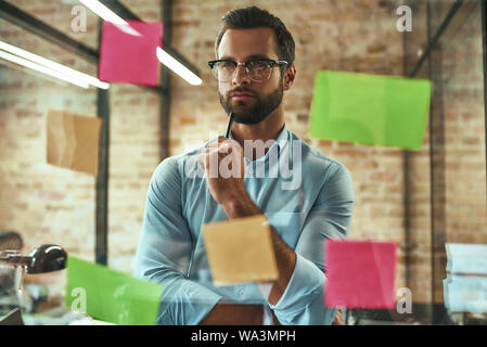 Startup. Young and handsome businessman in eyeglasses planning working process and using colorful stickers while standing in front of glass wall. Project management Stock Photo