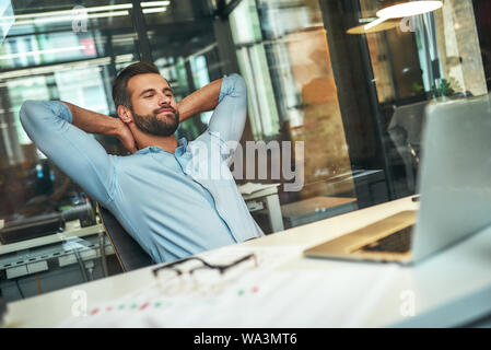 Work done Satisfied young bearded businessman leaning back with hands behind head and smiling while sitting in the modern office. Workplace. Taking a break Stock Photo