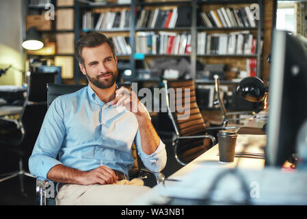 Confident and smart. Young bearded businessman in formal wear thinking about something and smiling while sitting in modern office. Working place. Business concept Stock Photo