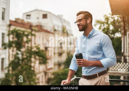 Young and handsome bearded man in eyeglasses and headphones holding cup of coffee and talking with friend while standing at the office balcony Stock Photo