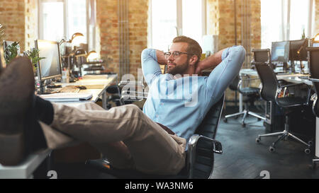 Work done. Side view of satisfied bearded young man in eyeglasses and formal wear holding hand behind his head and keeping legs on table. Modern office. Business concept Stock Photo