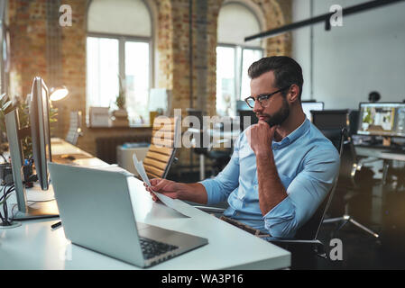 Analyzing data. Side view of concentrated bearded man in eyeglasses and formal wear looking at documents and touching his chin while sitting in the modern office. Business concept. Work concept Stock Photo