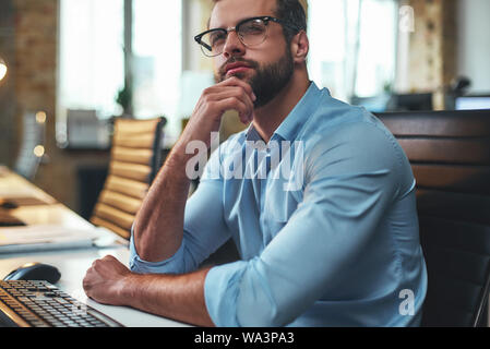 Time to think. Portrait of young bearded man in eyeglasses and formal wear touching his chin and looking away while working in the modern office. Business concept. Work concept. Modern technologies Stock Photo