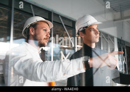 Building industry. Young engineer discussing with building worker on construction site. Industrial engineers have meeting while uses blueprint Stock Photo