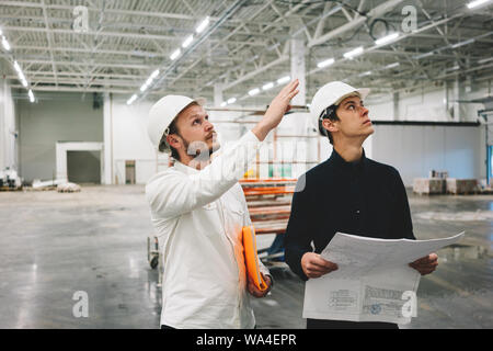 Building engineer and worker hold blueprint in hand while discussong construction site. Industrial engineers have meeting on build Stock Photo