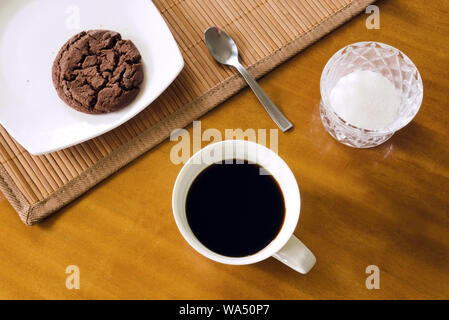 Black coffee in white cup, cookie on white plate, teaspoon, placemat and sugar-bowl on table, messy arrangement, top view Stock Photo